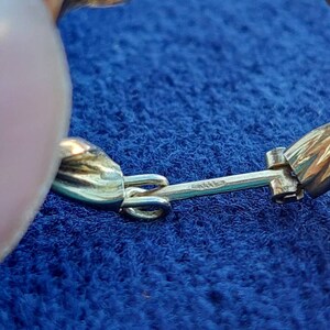 Puede incluir: Primer plano de un pendiente dorado con un dise&ntilde;o trenzado y un cierre de bisagra. El pendiente est&aacute; sobre una superficie de tela azul.