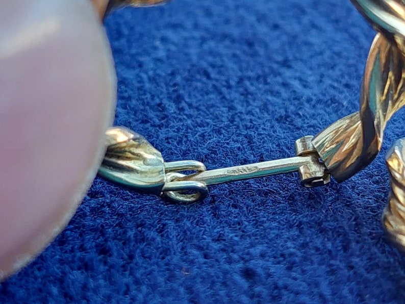 Puede incluir: Primer plano de un pendiente dorado con un dise&ntilde;o trenzado y un cierre de bisagra. El pendiente est&aacute; sobre una superficie de tela azul.