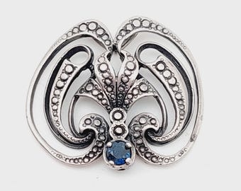 May include: A silver-toned brooch with an ornate, symmetrical design. The brooch features a central blue gemstone and is embellished with small, circular details. The overall shape is reminiscent of a stylized floral motif.