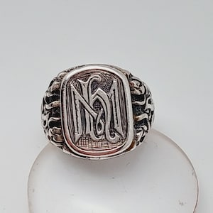 May include: A silver ring with a detailed design featuring the letters "NM" in a stylized font.