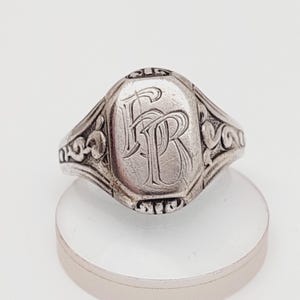 Op de afbeelding: Een zilveren zegelring met een gegraveerd monogram van de letters "FR". De ring heeft een decoratieve band met een bloemmotief.