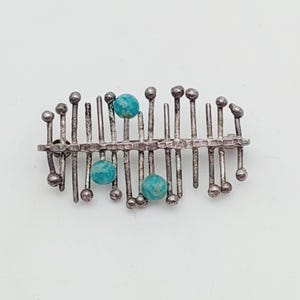 May include: A silver brooch with a linear design. The brooch features vertical posts with rounded tops, connected by a horizontal bar. Three turquoise beads are interspersed along the design. The brooch has a textured, handcrafted appearance.