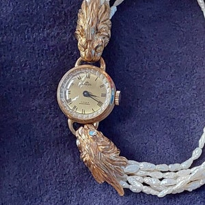 May include: A gold wristwatch with a white face and black hands. The watch is attached to a pearl necklace with two lion head clasps.