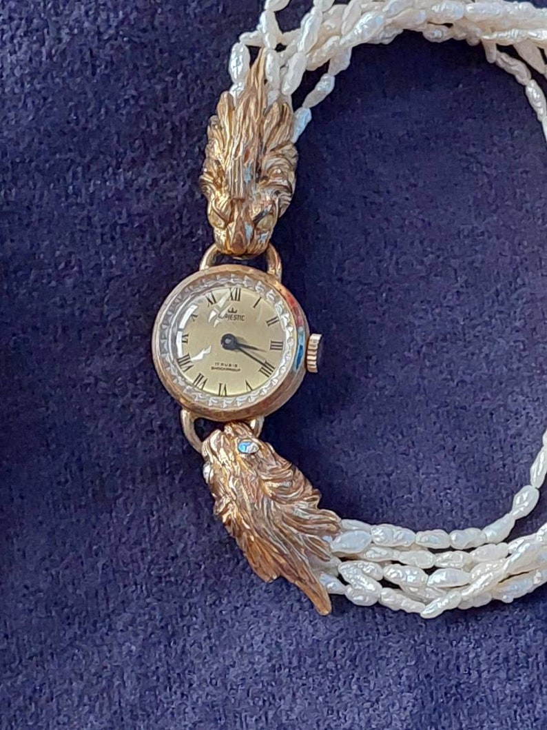 May include: A gold wristwatch with a white face and black hands. The watch is attached to a pearl necklace with two lion head clasps.