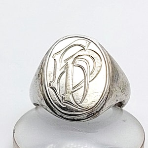 Peut inclure: Une chevalière en argent avec une face ovale. La face présente un monogramme gravé des lettres "DP" dans une police ornée et stylisée. La bague a une apparence classique et légèrement usée.