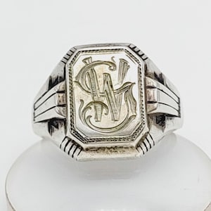 May include: A silver signet ring with an engraved monogram. The ring has a rectangular face with the initials "CW" and "M" in an ornate script. The band features a series of parallel lines on each side.