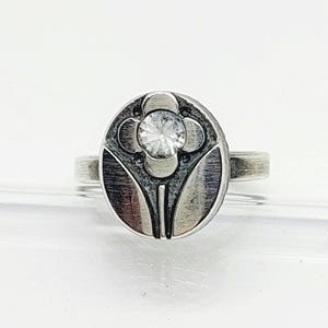Puede incluir: Anillo de plata con un diseño circular que presenta una piedra preciosa transparente central y un motivo floral. El anillo tiene un aspecto detallado y texturizado, con una banda que se estrecha hacia la parte posterior. El diseño recuerda al estilo Art Nouveau.