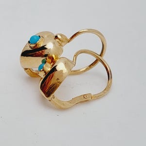 May include: A pair of gold-colored hoop earrings with turquoise-colored stone accents. The earrings have a unique design with a rounded shape and a hinged closure. The turquoise stones are small and round, adding a pop of color to the gold. The earrings are likely made of metal.