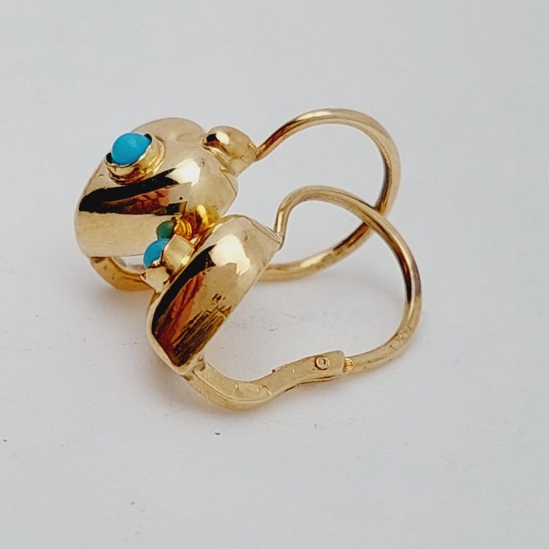May include: A pair of gold-colored hoop earrings with turquoise-colored stone accents. The earrings have a unique design with a rounded shape and a hinged closure. The turquoise stones are small and round, adding a pop of color to the gold. The earrings are likely made of metal.