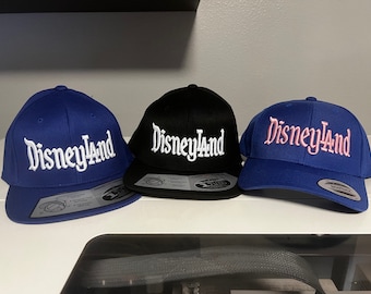 Könnte beinhalten: Drei Baseballkappen mit dem Wort "Disneyland" auf der Vorderseite gestickt. Die Kappen sind blau, schwarz und blau mit rosa Schrift.