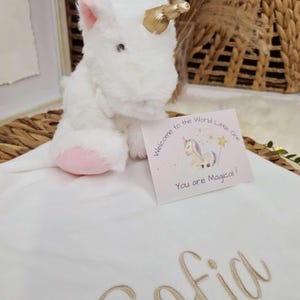 May include: A white plush unicorn with a gold horn and pink accents sits on a white blanket embroidered with the name "Sofia" in gold. A card reads "Welcome to the World Little One" and "You are Magical!"