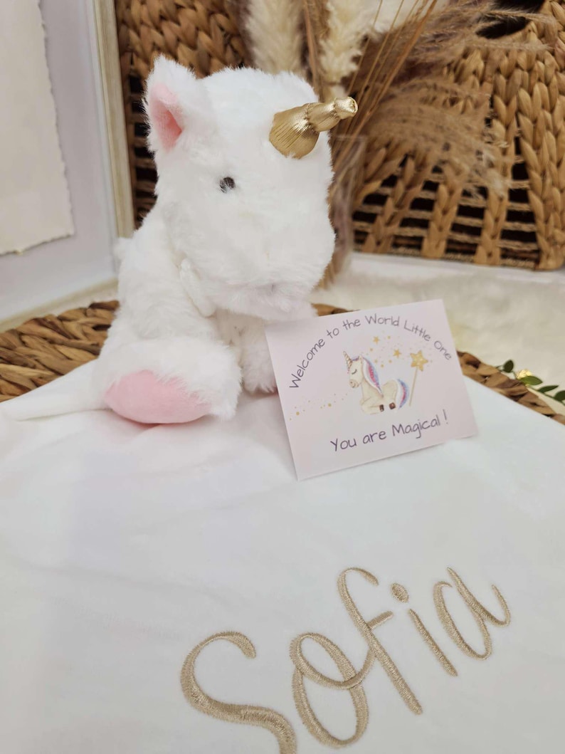 May include: A white plush unicorn with a gold horn and pink accents sits on a white blanket embroidered with the name "Sofia" in gold. A card reads "Welcome to the World Little One" and "You are Magical!"