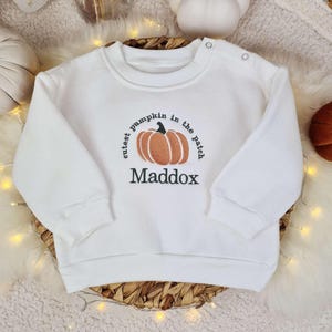 May include: White baby sweatshirt featuring the text "cutest pumpkin in the patch" and the name "Maddox" beneath a pumpkin graphic. The long-sleeved top has two shoulder poppers and is displayed on a woven basket.