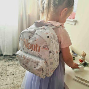 May include: A light gray backpack with a white zipper and the name "Noah" embroidered in orange. The backpack features a pattern of gray koala bears and pink accents. The child is wearing a pink shirt and a light purple skirt.