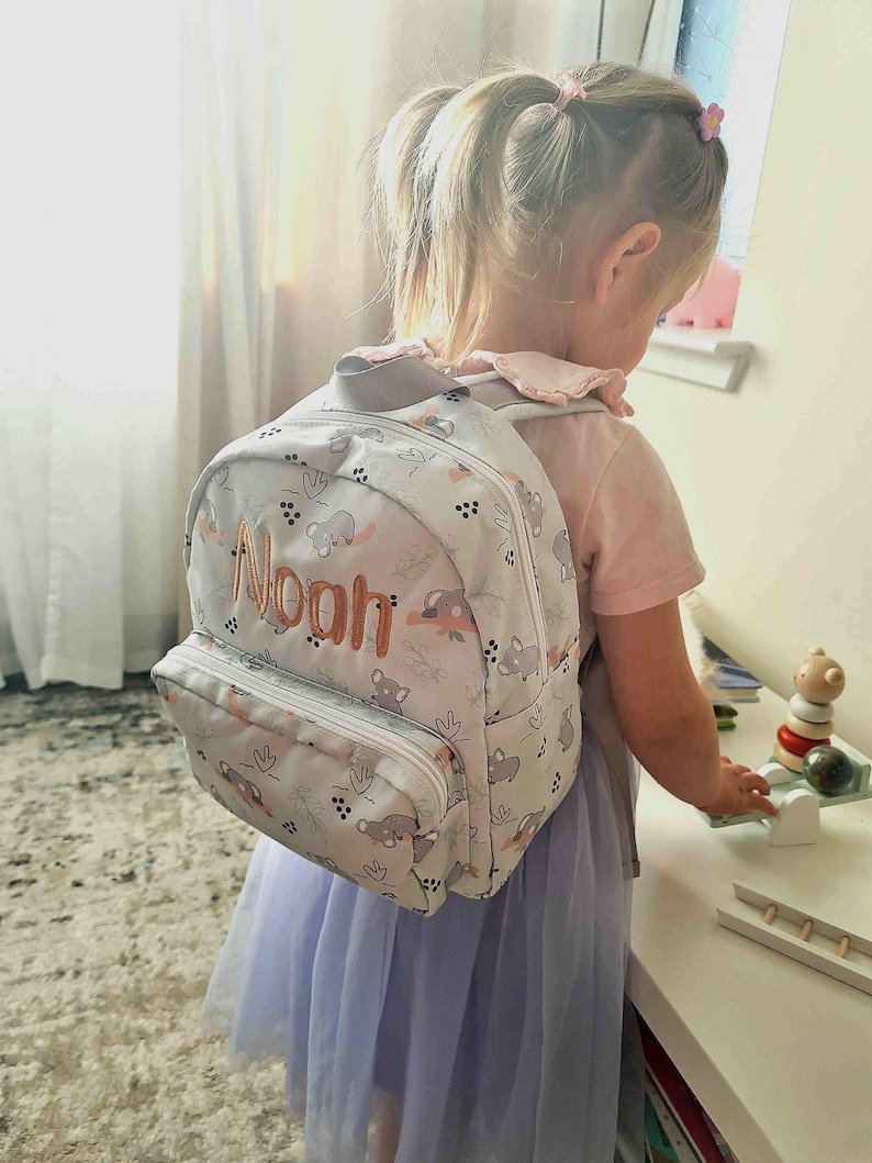 May include: A light gray backpack with a white zipper and the name "Noah" embroidered in orange. The backpack features a pattern of gray koala bears and pink accents. The child is wearing a pink shirt and a light purple skirt.