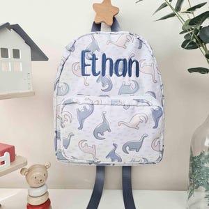 May include: A white backpack with a pattern of blue and pink dinosaurs. The name "Ethan" is embroidered in blue on the front. The backpack has blue straps and is hanging from a wooden star.