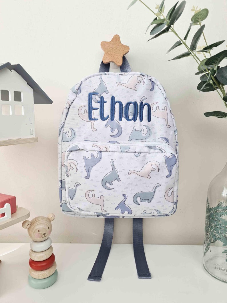 May include: A white backpack with a pattern of blue and pink dinosaurs. The name "Ethan" is embroidered in blue on the front. The backpack has blue straps and is hanging from a wooden star.