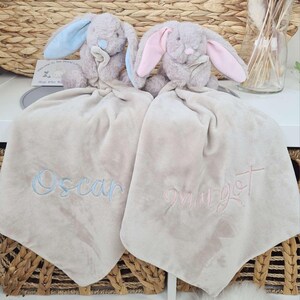 May include: Two plush bunny security blankets. One bunny has blue ears, the other has pink ears. Each blanket is beige and embroidered with a name in a matching color. A card reads "Welcome to the world little one."