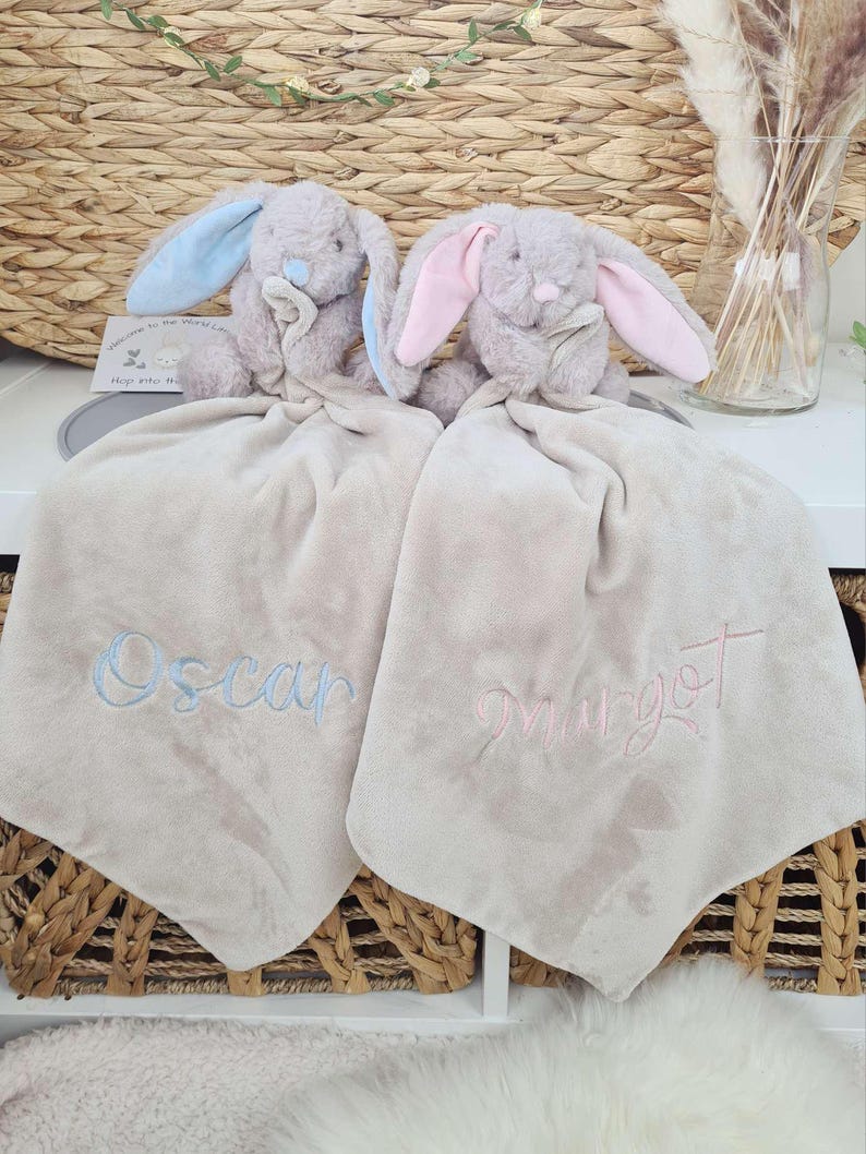 May include: Two plush bunny security blankets. One bunny has blue ears, the other has pink ears. Each blanket is beige and embroidered with a name in a matching color. A card reads "Welcome to the world little one."