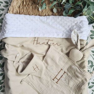 May include: A beige muslin swaddle blanket with the name "Hunter" embroidered on it. The blanket is folded in half and has a white plush lining. There is a matching beige muslin burp cloth with the letter "H" embroidered on it. There is also a beige muslin bunny toy with a floral print on the ear.