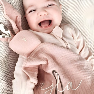 May include: A baby in a light pink onesie smiles, holding a pink bunny comforter. The comforter features the name "Ajla" embroidered in white script. The bunny has pink ears and a white triangle nose. The baby is lying on a patterned blanket.