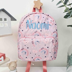May include: Pink backpack with a rainbow and cloud pattern, featuring the name "Naomi" embroidered in blue. The backpack has pink straps and a wooden star-shaped hanger.