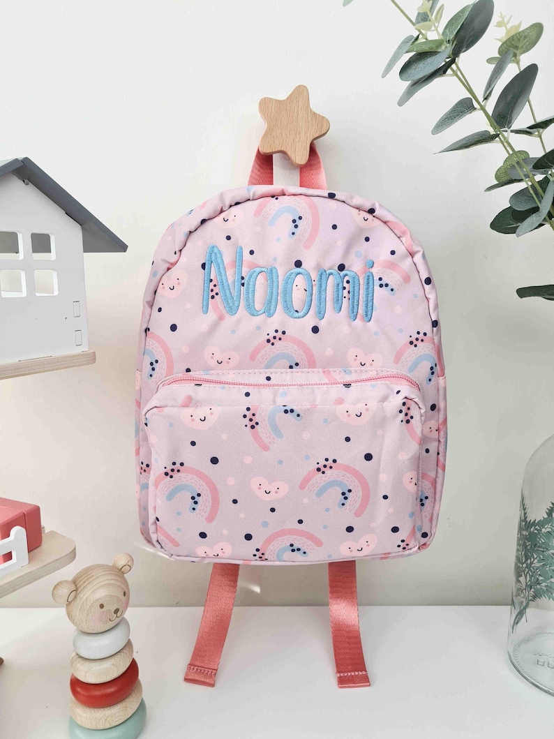 May include: Pink backpack with a rainbow and cloud pattern, featuring the name "Naomi" embroidered in blue. The backpack has pink straps and a wooden star-shaped hanger.