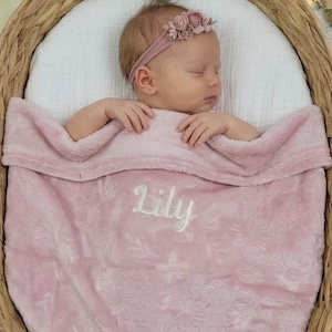 May include: A sleeping baby in a woven basket, covered with a pink blanket embroidered with "Lily." The baby wears a floral headband. The blanket has a floral design. The basket is wicker.