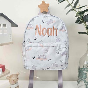 May include: A light blue backpack with a pattern of elephants and leaves, featuring the name "Noah" in embroidered orange thread. The backpack has gray straps and a small front pocket. It hangs from a wooden star-shaped hook.