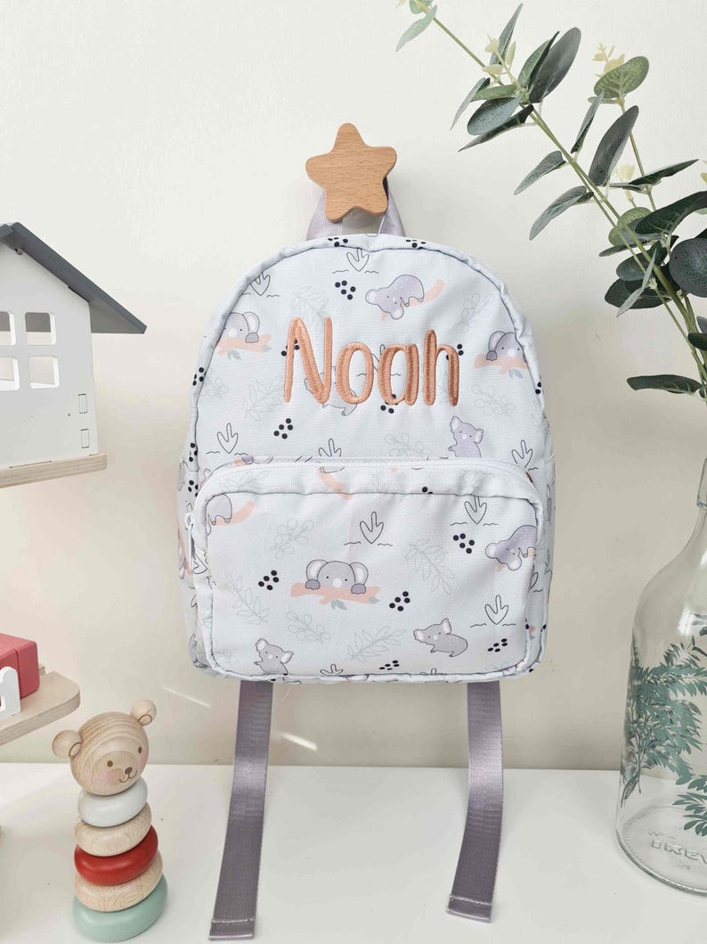 May include: A light blue backpack with a pattern of elephants and leaves, featuring the name "Noah" in embroidered orange thread. The backpack has gray straps and a small front pocket. It hangs from a wooden star-shaped hook.