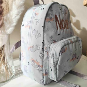 May include: A light gray backpack with a koala bear pattern and the name "Noan" embroidered in brown. The backpack has a front pocket with a zipper, gray straps, and a top handle. The koala pattern includes pink and gray accents.