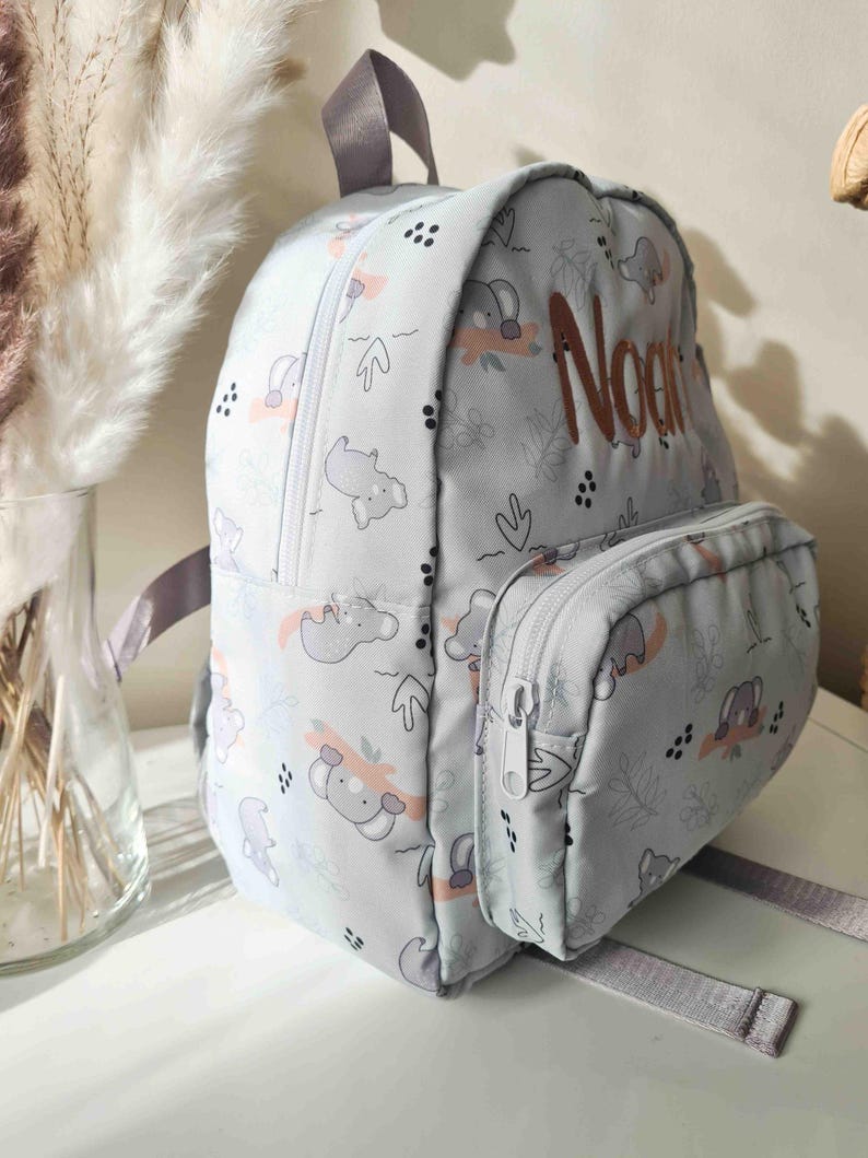 May include: A light gray backpack with a koala bear pattern and the name "Noan" embroidered in brown. The backpack has a front pocket with a zipper, gray straps, and a top handle. The koala pattern includes pink and gray accents.
