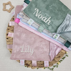 May include: A selection of baby comfort blankets in a woven basket. The blankets are in pastel shades of pink, blue, and green, with embroidered names and floral designs. Each blanket has colourful ribbons and tags attached.