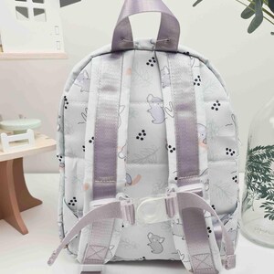 May include: A small, light gray backpack with a repeating pattern of cartoon animals and floral elements. The backpack has lavender straps and a top handle. The straps are adjustable with a clear plastic buckle. The backpack is designed for children.