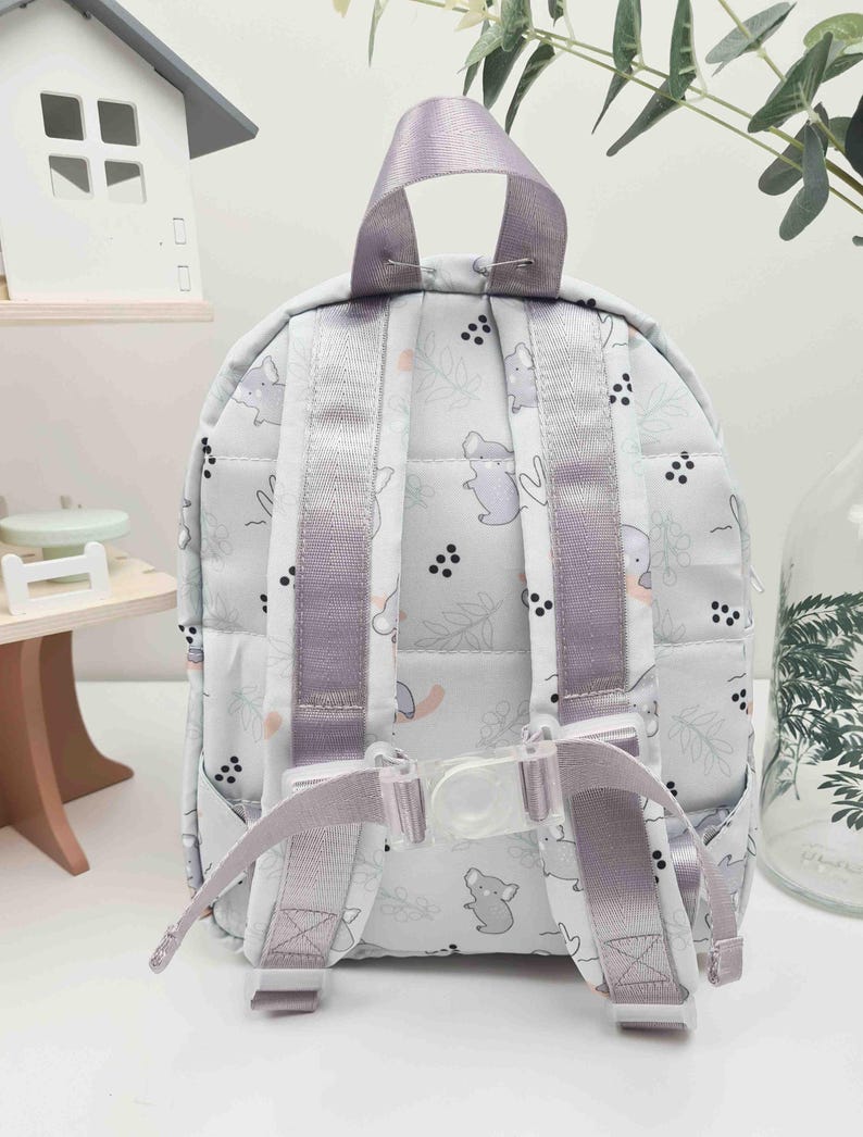 May include: A small, light gray backpack with a repeating pattern of cartoon animals and floral elements. The backpack has lavender straps and a top handle. The straps are adjustable with a clear plastic buckle. The backpack is designed for children.