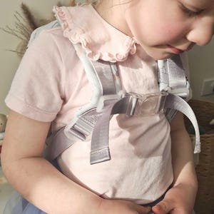 May include: A child wearing a light gray baby carrier with adjustable straps and a clear plastic buckle. The carrier has a light blue and white patterned design. The child is wearing a pink shirt with a ruffled collar and a light blue skirt.