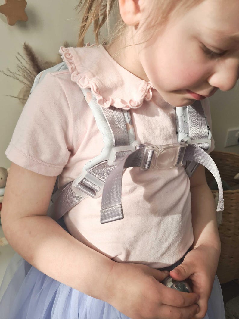 May include: A child wearing a light gray baby carrier with adjustable straps and a clear plastic buckle. The carrier has a light blue and white patterned design. The child is wearing a pink shirt with a ruffled collar and a light blue skirt.
