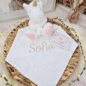May include: A white baby blanket with the name "Sofia" embroidered in gold thread. A plush white unicorn with a gold horn and pink accents sits atop the blanket. A card reads "Welcome to the World Little One, You are Magical!"