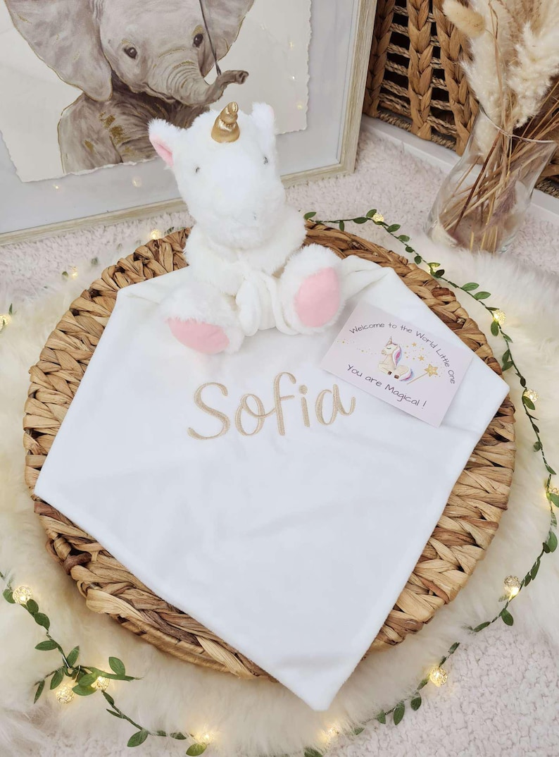 May include: A white baby blanket with the name "Sofia" embroidered in gold thread. A plush white unicorn with a gold horn and pink accents sits atop the blanket. A card reads "Welcome to the World Little One, You are Magical!"