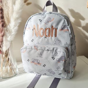 May include: A light gray backpack with a repeating pattern of gray elephants and coral-colored accents. The name "Noah" is embroidered in a copper color on the front. The backpack has a front pocket, gray straps, and a top handle.