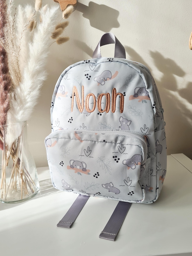 May include: A light gray backpack with a repeating pattern of gray elephants and coral-colored accents. The name "Noah" is embroidered in a copper color on the front. The backpack has a front pocket, gray straps, and a top handle.