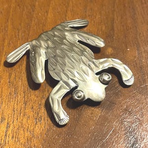 May include: A silver-toned metal frog-shaped brooch. The brooch has detailed textures on its body and legs, with two round eye details. The frog is positioned on a wooden surface.
