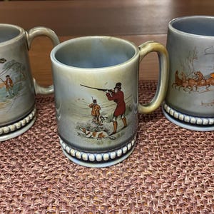 May include: Three ceramic mugs with detailed hunting scenes. Each mug has a light green-gray base color, a cream-colored handle, and a decorative base. The scenes depict figures with hunting dogs and a horse-drawn carriage.