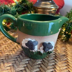 May include: A green ceramic pitcher with a brown rim and two white sheep with black faces on the front. The pitcher has a handle and a spout.
