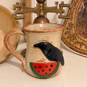 May include: Handmade ceramic mug with a raven and watermelon design. The mug features a tan base with a brown handle and rim. A black raven is perched above a red watermelon slice with black seeds. The mug is a unique piece of kitchenware.
