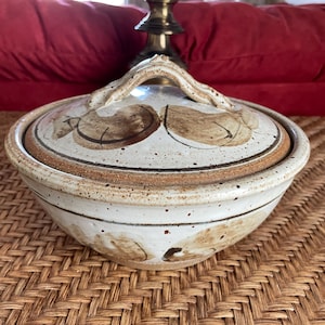 May include: A white ceramic casserole dish with a brown lid and a unique handle. The dish has a speckled glaze and brown leaf-like designs.