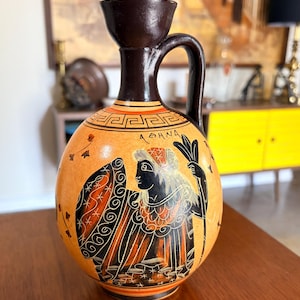 May include: A vintage, round-bodied ceramic vase with a dark brown handle and rim. The vase features a painted design of a figure with wings, holding a sprig, in black, orange, and white against a tan background. The word "ATHNA" is written on the vase.