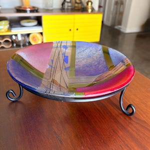 May include: A colorful glass bowl with a metal stand. The bowl features a mix of red, purple, blue, and green hues in a geometric pattern. The metal stand has a dark finish and a curved design.