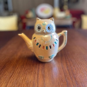 May include: A ceramic teapot shaped like an owl with blue eyes and a colorful design around the neck. The teapot is a light brown color.