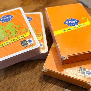 May include: Orange Etap Hotel playing cards and card boxes. The cards and boxes feature the Etap Hotel logo, the phrase "Au cœur du Tour!" and images of cyclists. The cards are stacked on a wooden surface.